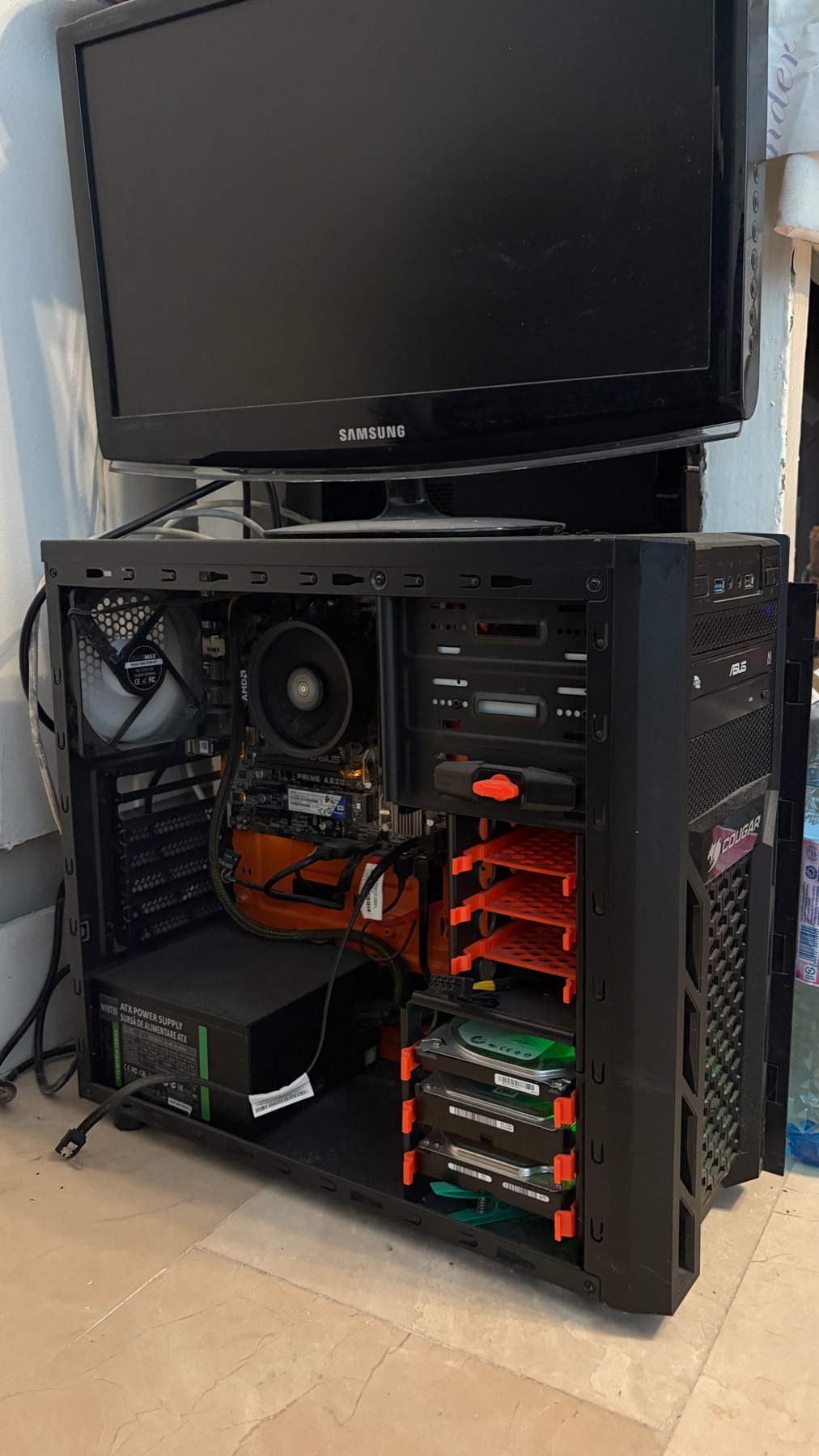 The TrueNAS tower with its side panel off: Ryzen cooler, ASUS PRIME A320 board and three hard drives in orange caddies, an old Samsung monitor sitting on top
